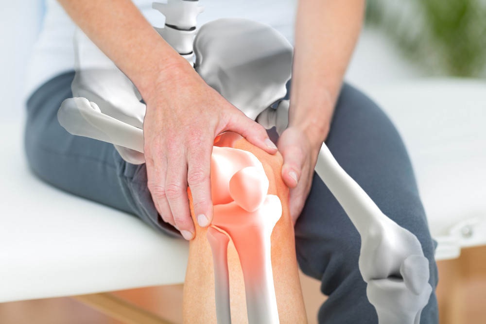 Zdjęcie przedstawiające mężczyznę trzymającego się za kolano. Na zdjęciu widać jakby otwartą nogę i kości. Na łączeniu w kolanie jest czerwony obrzęk sygnalizujący ból.